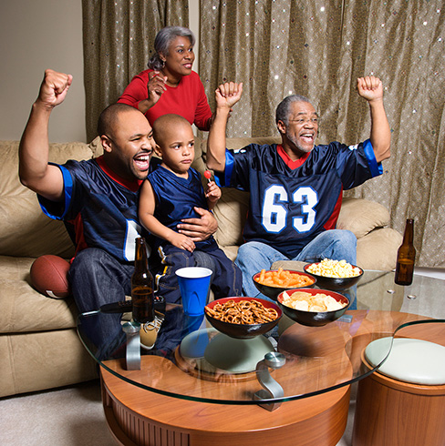 Family of four all wearing sports jerseys, cheering on a gootball game from their couch.